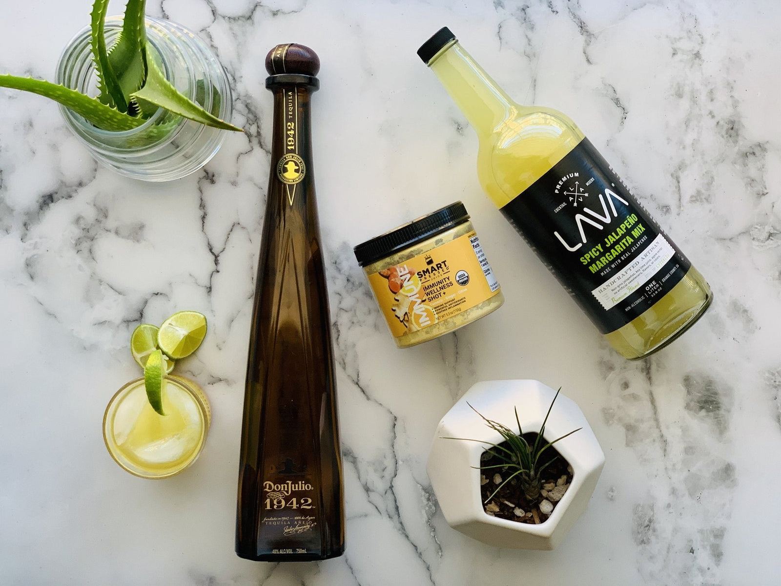 1 glass of CJ's Immunity Margarita beside 1 bottle of tequila beside a jar of Immunity Wellness beside a bottle of Margarita mix side by side. On top is an aloe vera plant and on the bottom is another plant.