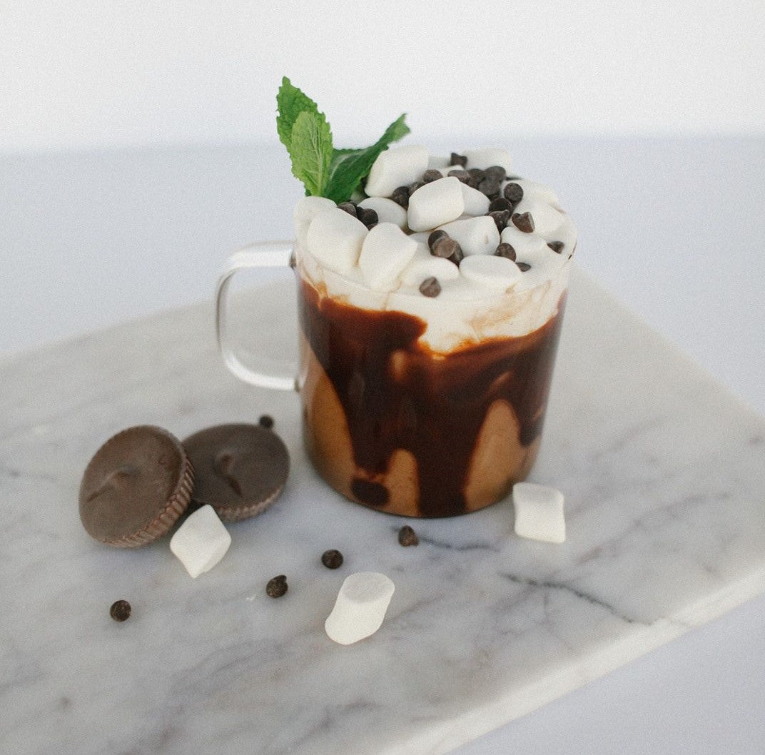 side view of a mug with proteini hot chocolate topped with marshmallows and chocolate bits and mints with two circle-shaped chocolate, three mallows, and choco bits on top of a marble table