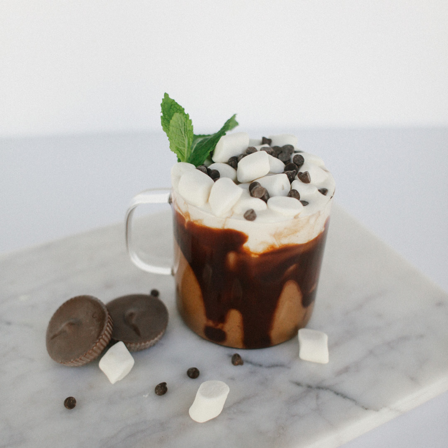 a cup of hot chocolate drink topped with marshmallow, chocolate chips, and a green leaf. On the surface are 2 round-shaped chocolates, marshmallows, and some chocolate chips.