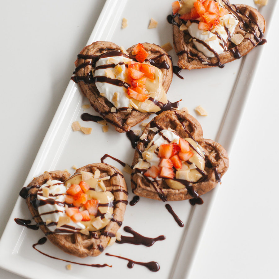 4 heart-shaped waffles topped with nut butter, whipping cream, and chopped mix fruits drizzled with chocolate syrup on a white rectangular plate.
