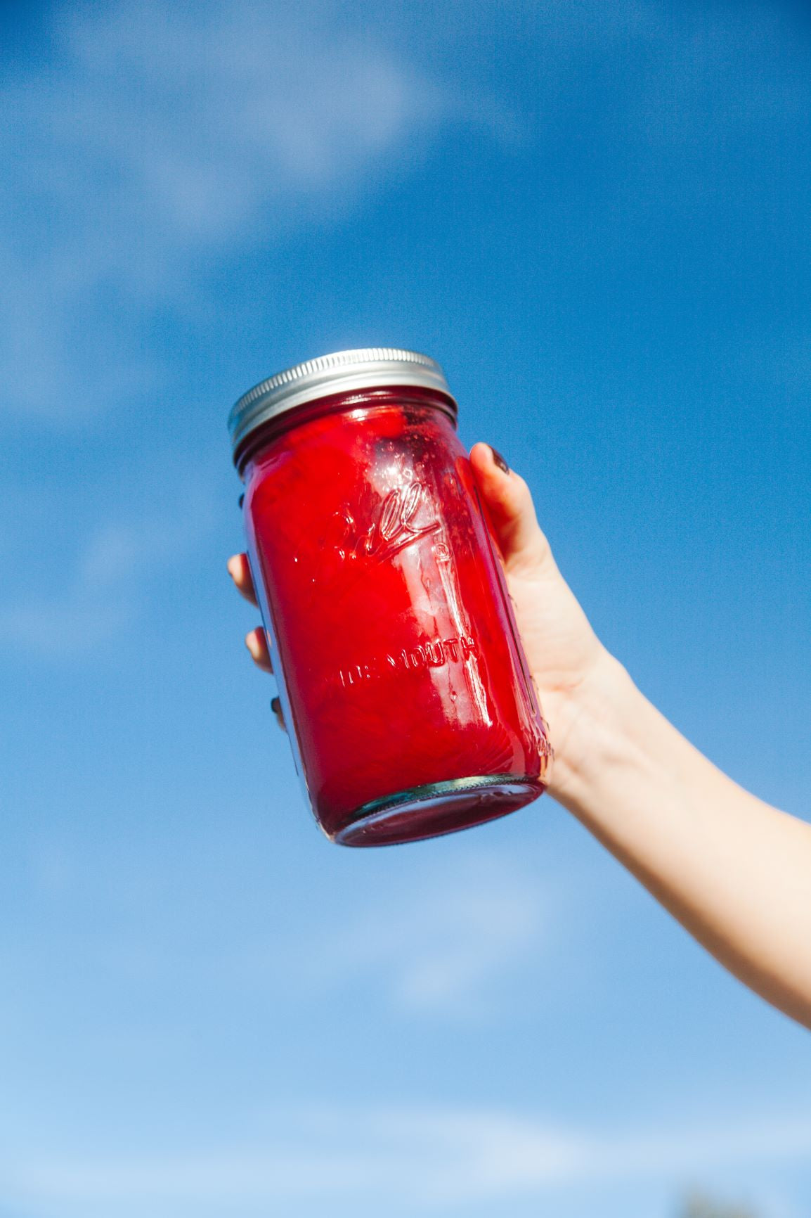 A hand of a woman holding a large mason jar filled with red juice with a blue sky as a background