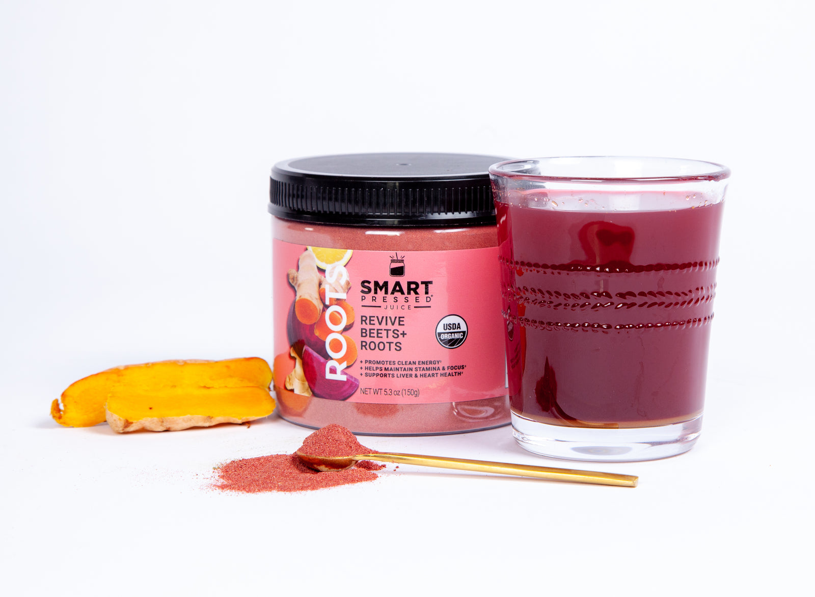 a jar of Revive Beet+Roots beside a glass of Revive Beet+Roots juice, on the surface is a spoon with pink powder and a slice of turmeric, Set against a white background.