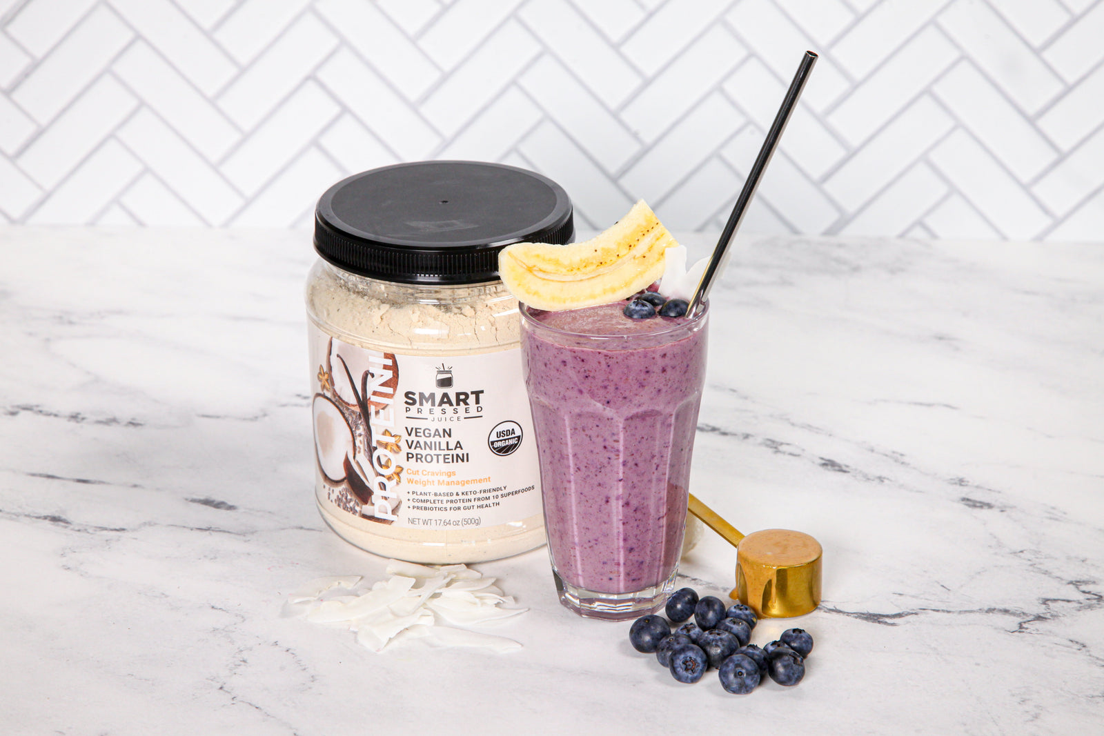 1 jar of Vegan Vanilla Proteini beside a glass of Trina's Blueberry Butter Proteini with a straw topped with a sliced banana and blueberries beside a golden scoop and blueberries on a marble counter top.