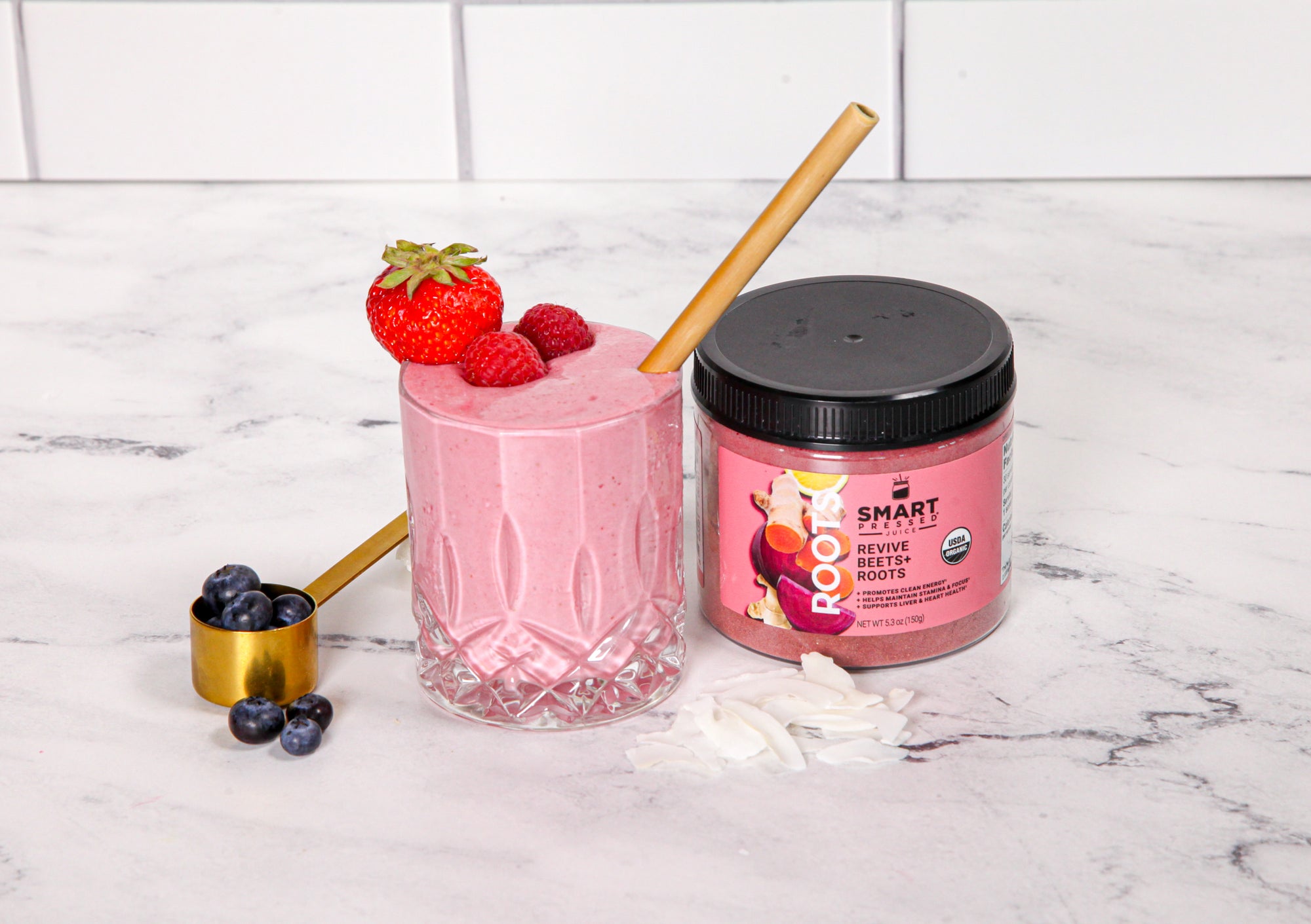 a glass of berry beets smoothie topped with strawberries with a brown paper straw and a smart revive beetroots labeled jar and a golden scoop filled with blueberries on the side on top of a marble table 