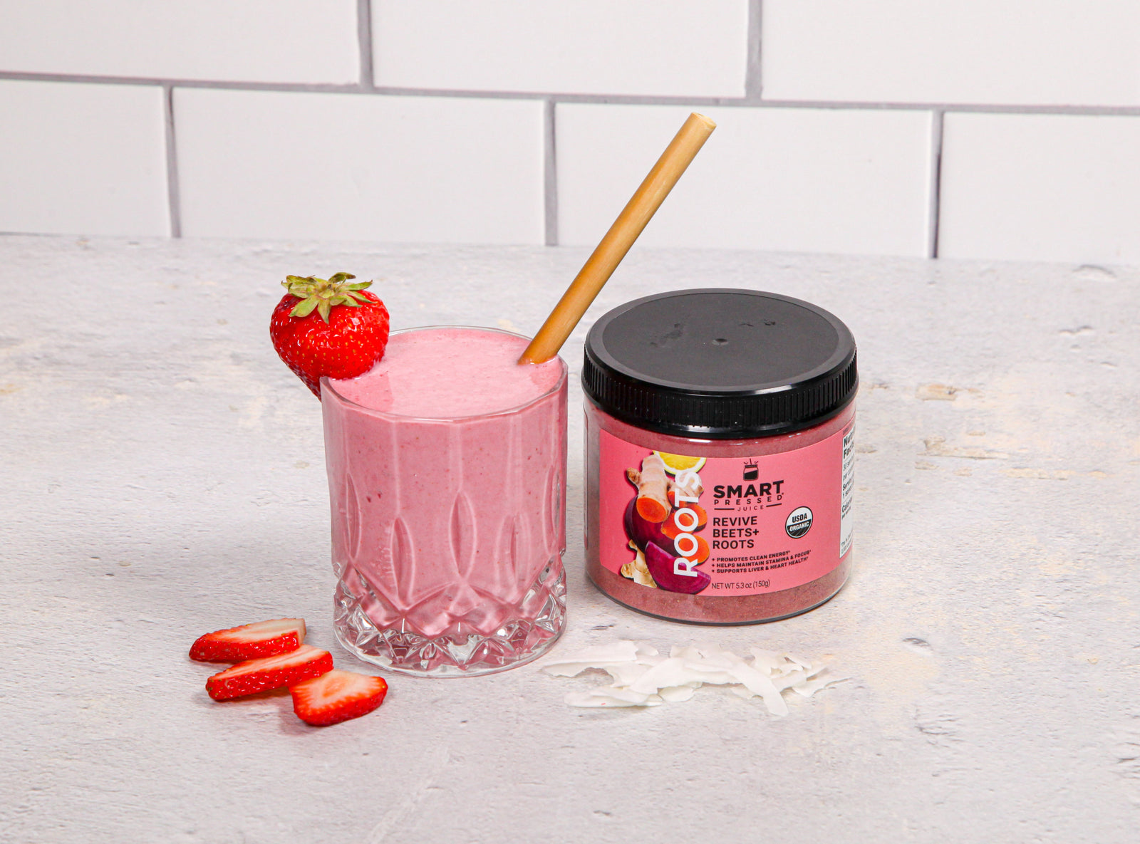 a glass of strawberry beets smoothie garnish with strawberry with a brown paper straw and smart beetroot labeled jar on the side and slices of strawberry on top of a white table