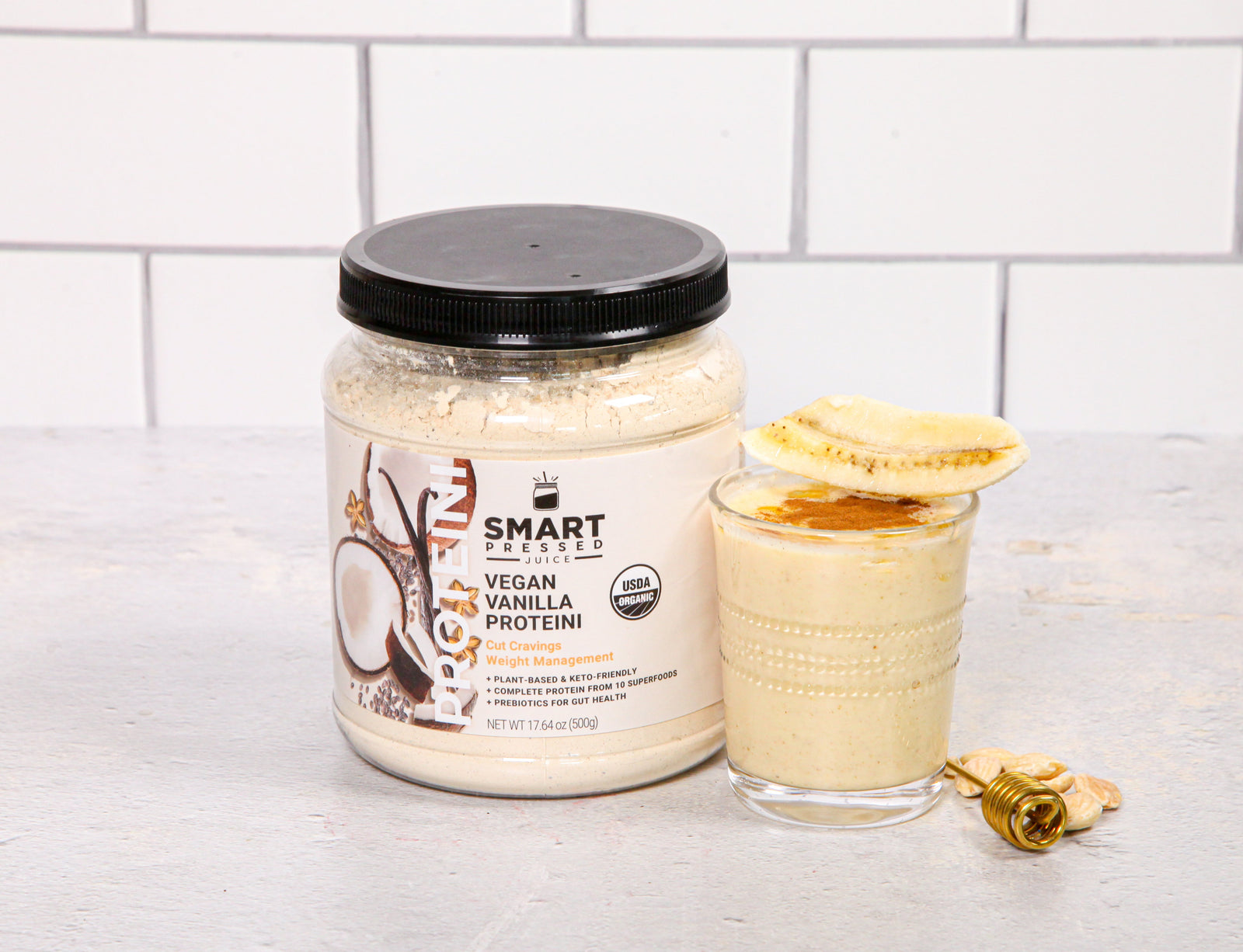 1 jar of Vegan Vanilla Proteini beside a glass of Amanda's PB & Honey Proteini topped with honey and a sliced banana beside a honey dipper on a marble counter top.