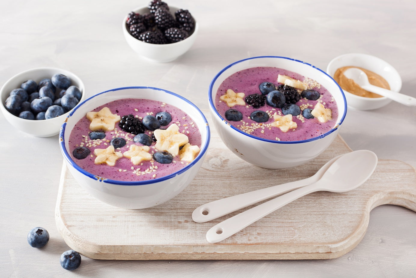 2 bowl of blue berry Vegan Vanilla Proteini smoothie topped with blue berries and star-shaped bananas beside 2 plastic spoon on a cutting board beside 1 bowl of blue berries, 1 bowl of black berries, and 1 dipping saucer of peanut butter with a spoon.