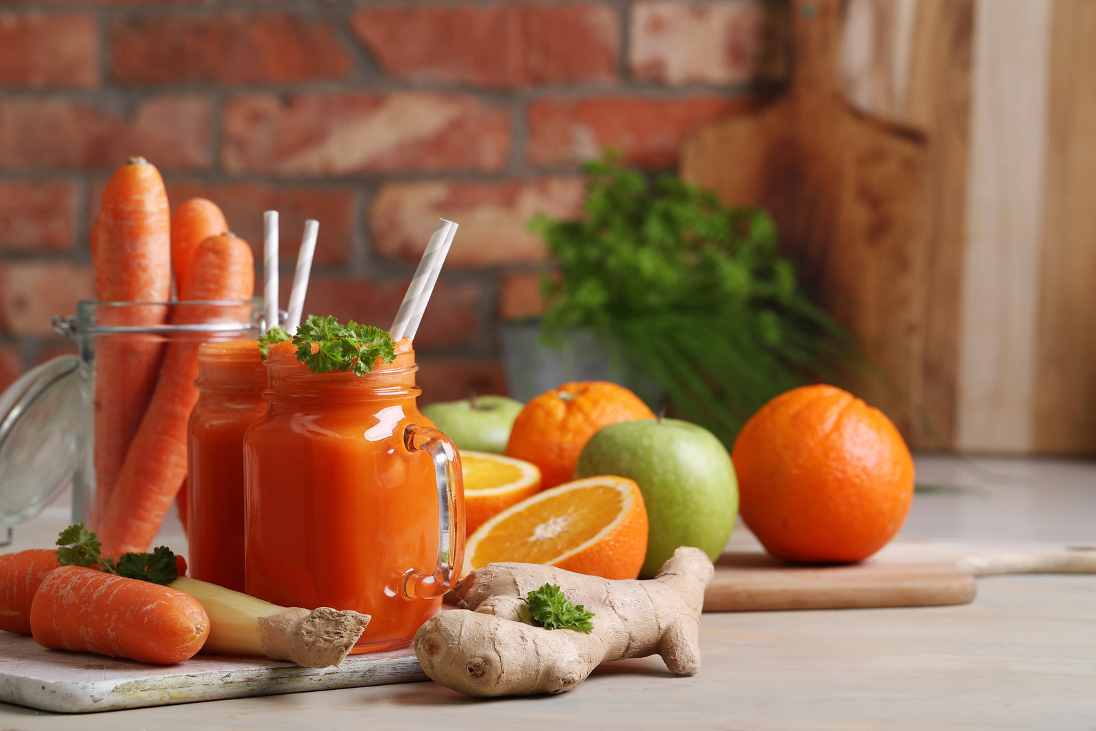 Image of 2 jars of orange-colored smoothie with a straw surrounded by 3 carrots in a jar, 1 carrot on the surface, a slice of ginger, half an orange, 2 oranges, and 2 green apples.