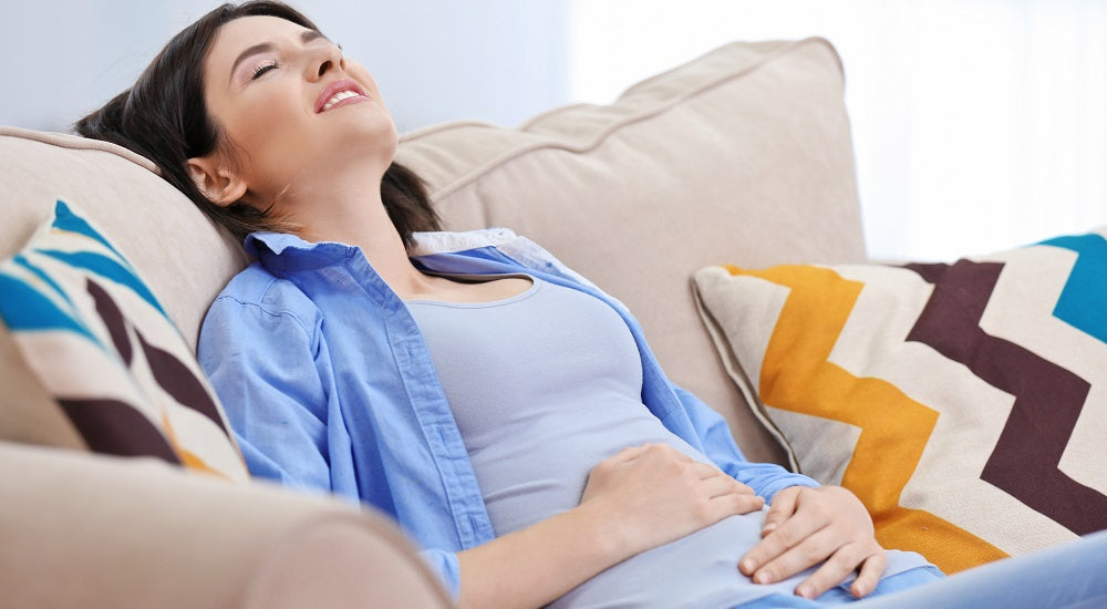 Woman lying back on beige sofa holding her stomach with striped pillows on either side of her