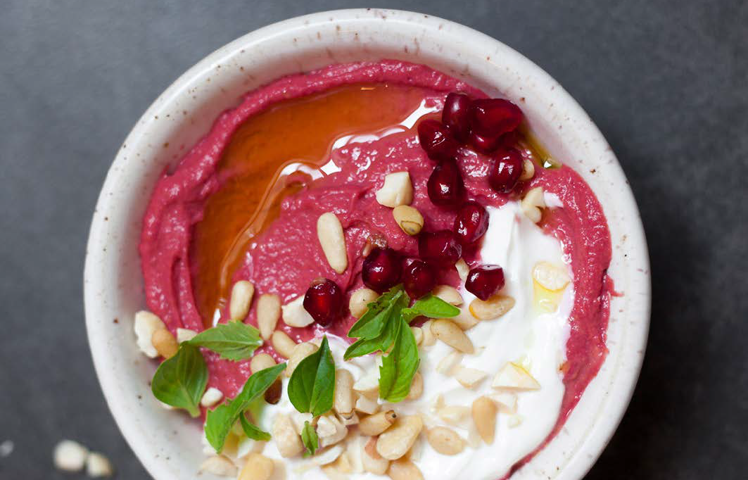 top view shot of a bowl of Revive Beet+Roots hummus topped with pomegranate seeds, coconut cream, sunflower seeds, olive oil, and green leaves.
