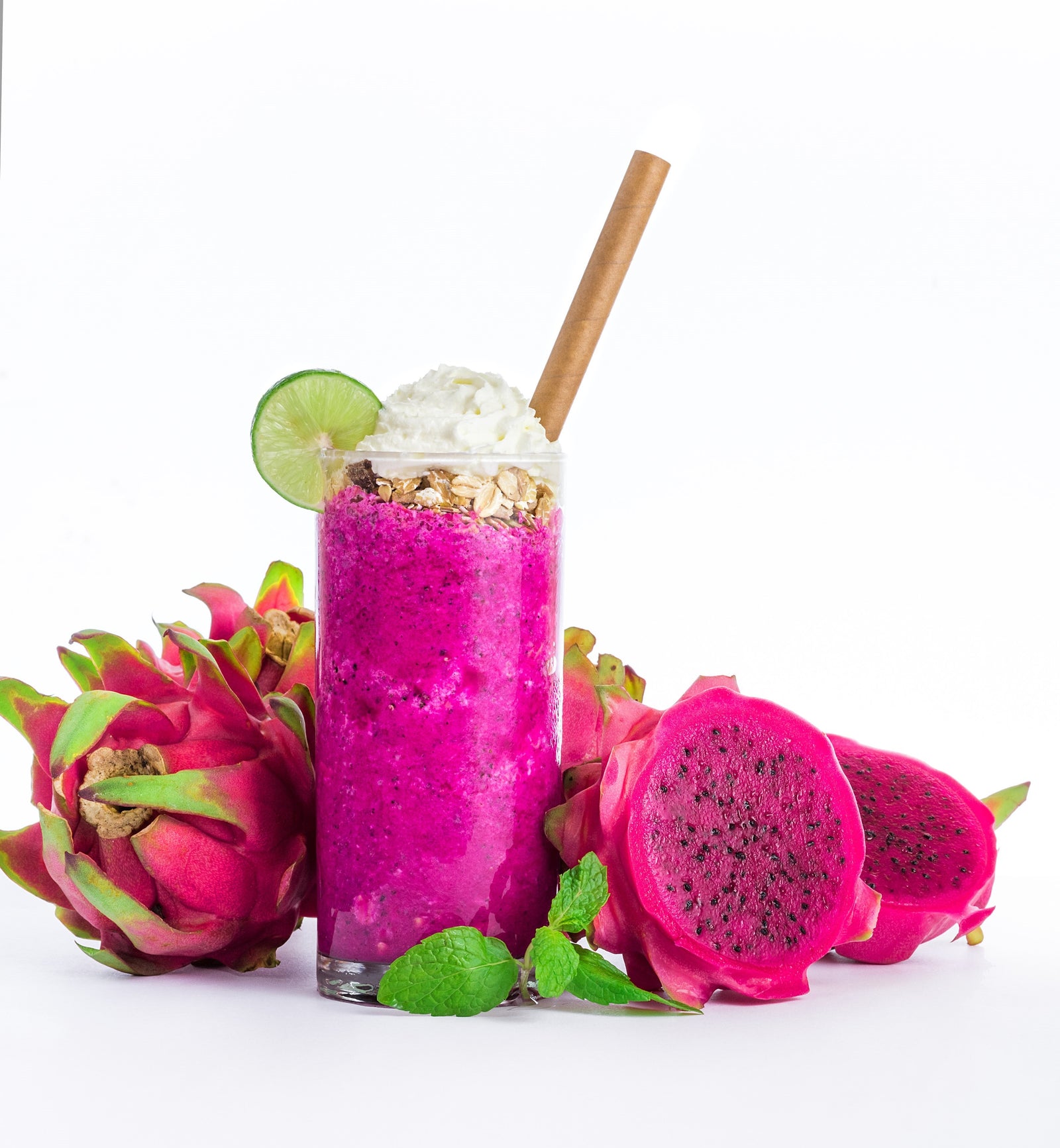 a glass of pineapple pitaya smoothie topped with whip cream, nuts, and a slice of lime with brown paper straw surrounded by half-sliced dragon fruit with mint leaves on the side and whole dragon fruit