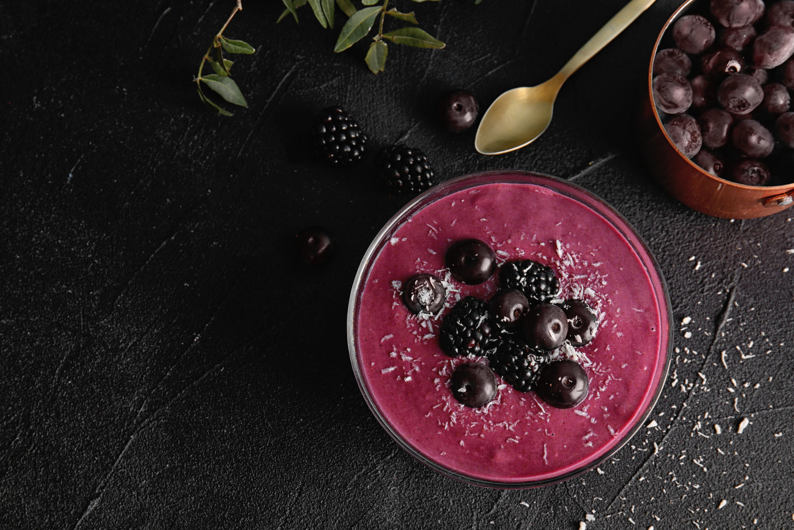 top view shot of blueberry smoothie in a bowl topped with blueberries, blackberries and coconut shreds beside a lid container of blueberries, a golden spoon, 1 blueberry, 2 blackberries, and leaves on a black surface.