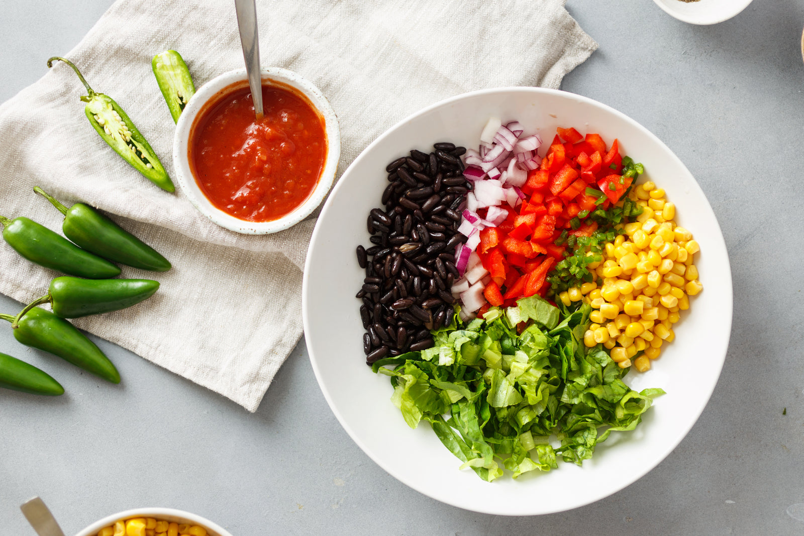 top view of a white ceramic plate with corn cob, sliced tomato, onion, black beans, and lettuce with a small white bowl with avocado jalapeno creme with a spoon on top of a white table cloth with five whole jalapenos and one half sliced jalapeno