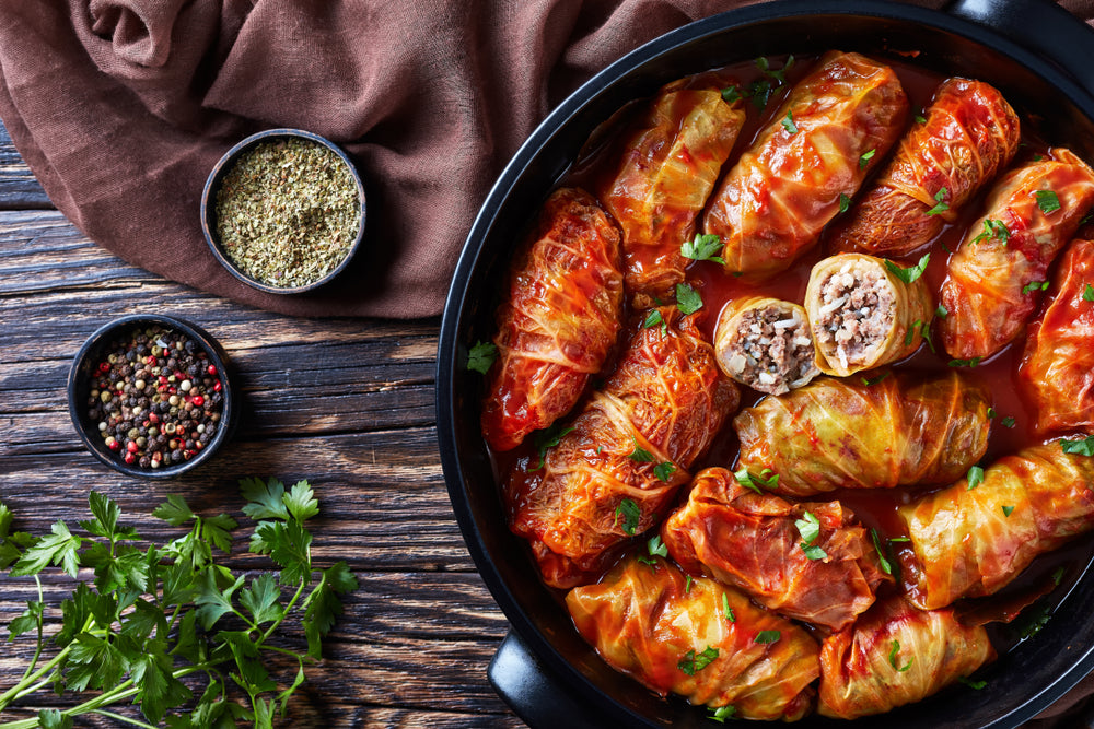 top view of barley stuffed cabbage rolls with brown cloth and small bowls with peppercorn and dried basil and parsley on the side on top of a wooden table.