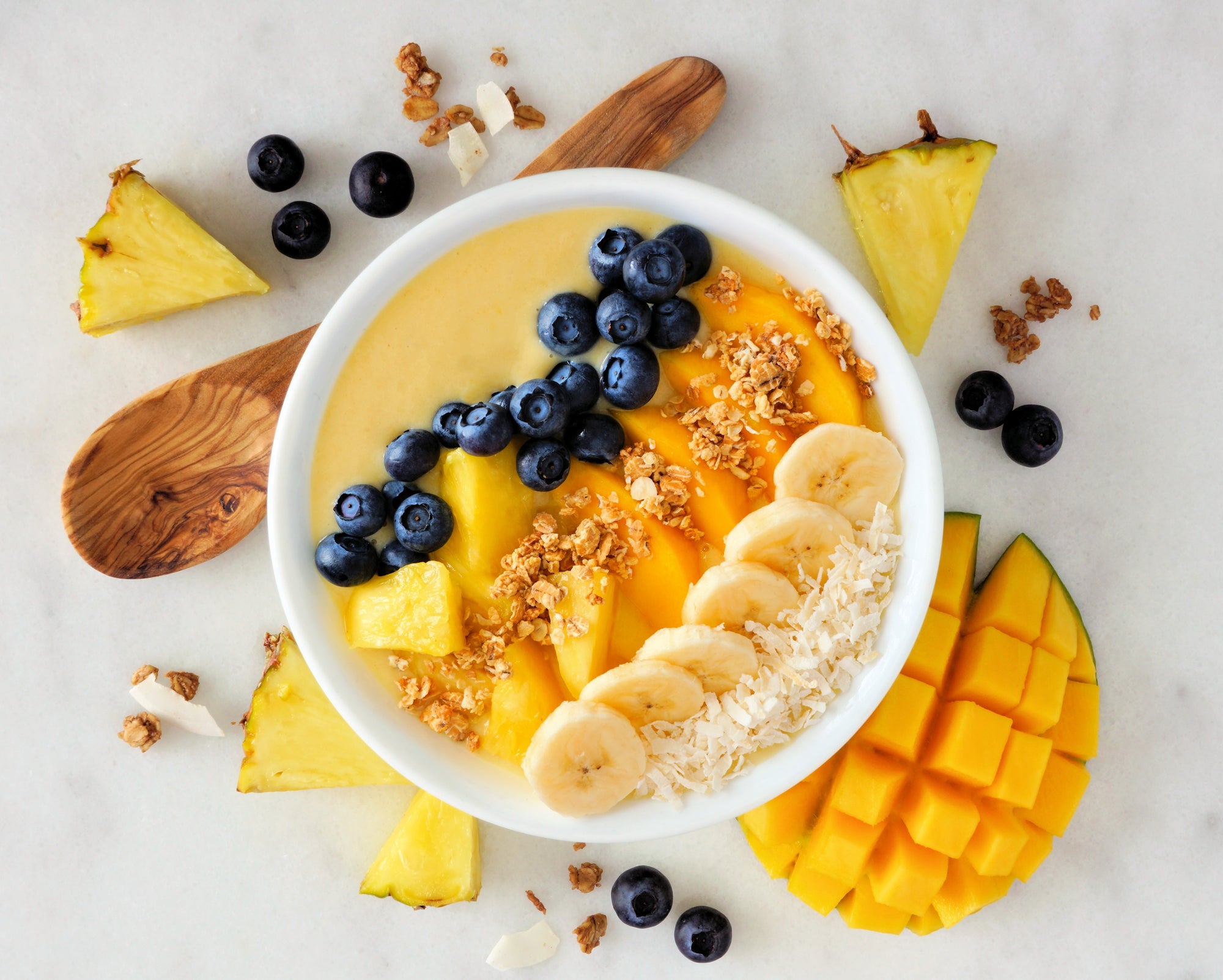 top view shot of a bowl of yellow smoothie topped with blueberries, slices of mango, banana, mango, and sprinkled with granola and coconut shreds. On the surface is a slice of ripe mango, blueberries, pineapple wedges, granola, and a wooden spatula.