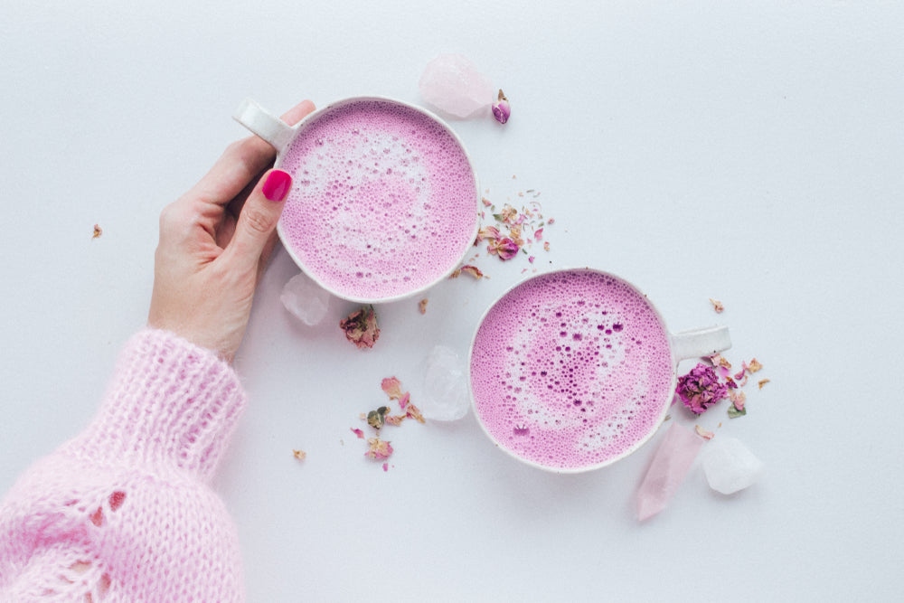 top view of two cups of pink beets latte with a hand holding a cup with scattered dried flowers on top of a white table