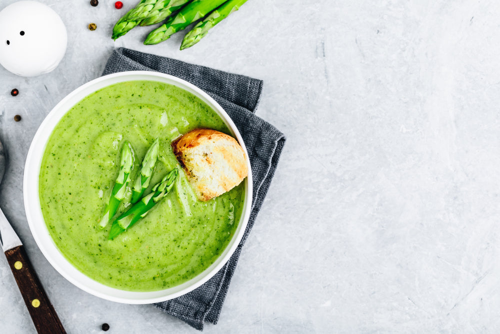 top view of a lemon asparagus soup topped with slices of asparagus on top of a black folded table cloth with a bread knife, fresh asparagus, peppercorns on the side