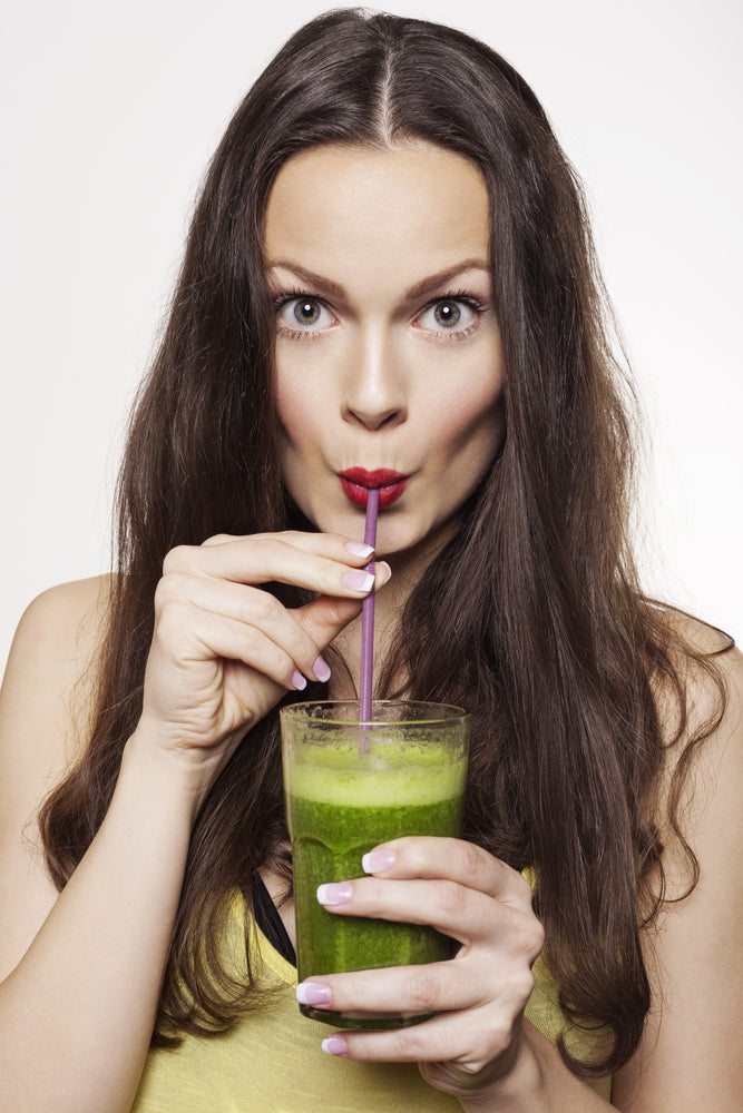 A long-haired woman wearing a yellow spaghetti strap holding a glass of green smoothie with her right hand holding a violet paper straw while sipping the green smoothie with her eyes wide open.