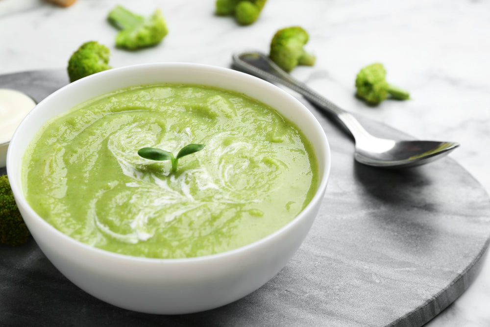 top view of a bowl with creamy broccoli soup with a spoon on the side on top of a circle-shaped wood with broccoli florets on top of a marble table