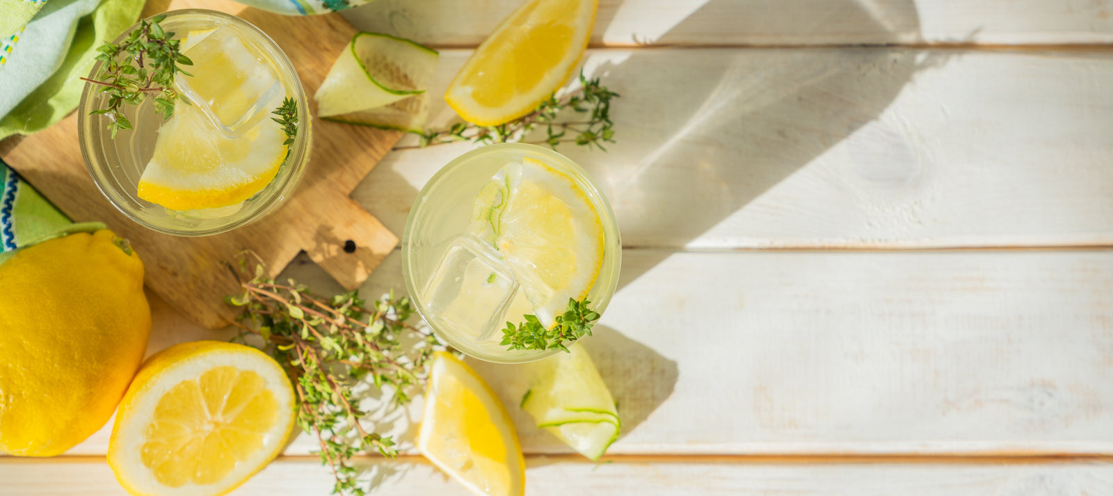 top view of two glasses of lemon juice with ice on top of white painted wooden table surrounded with one whole lemon and slices of lemon