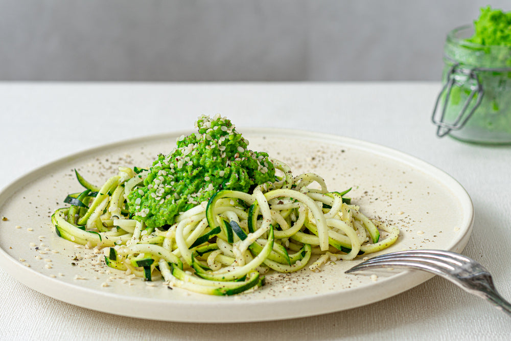 side view of a white ceramic plate with Pesto Zoodles with a fork on the side and a small jar of organic pressed greens on top of a white table