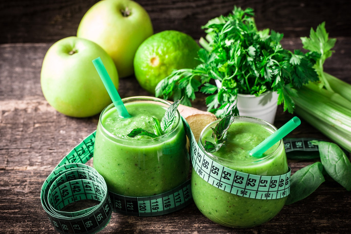 2 glasses of Organic Pressed Green smoothie with green straw wrapped with measuring tape. Behind is a celery, cilantro, 1 lime, and 2 green apples on a wooden surface.