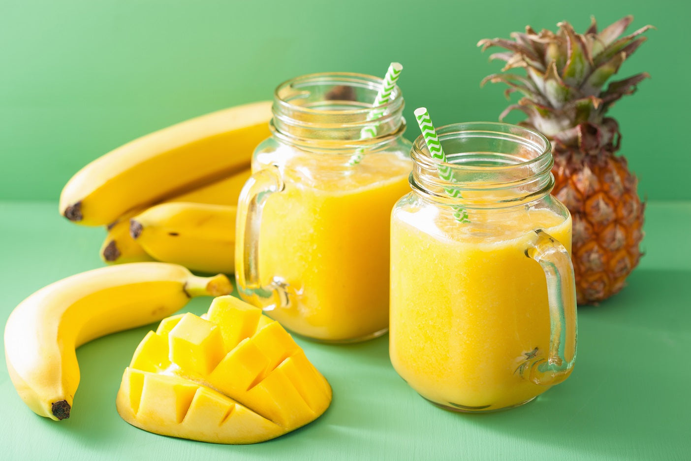 2 mason jars of Mango Banana Cleanse next to a pineapple, sliced mango, and bananas