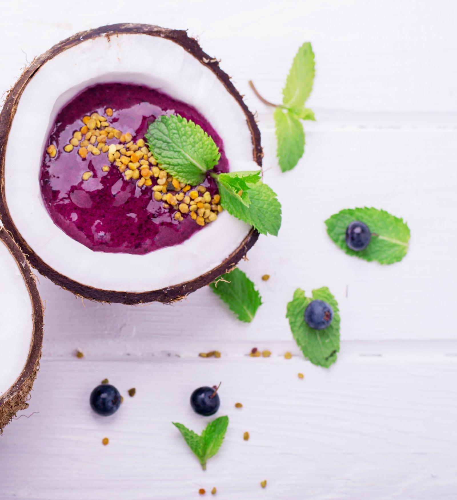 top view of a coconut cut in halves filled with coconut water pineapple smoothie topped with dried berries and mint surrounded by blueberries, dried berries, and mint on top of a white table