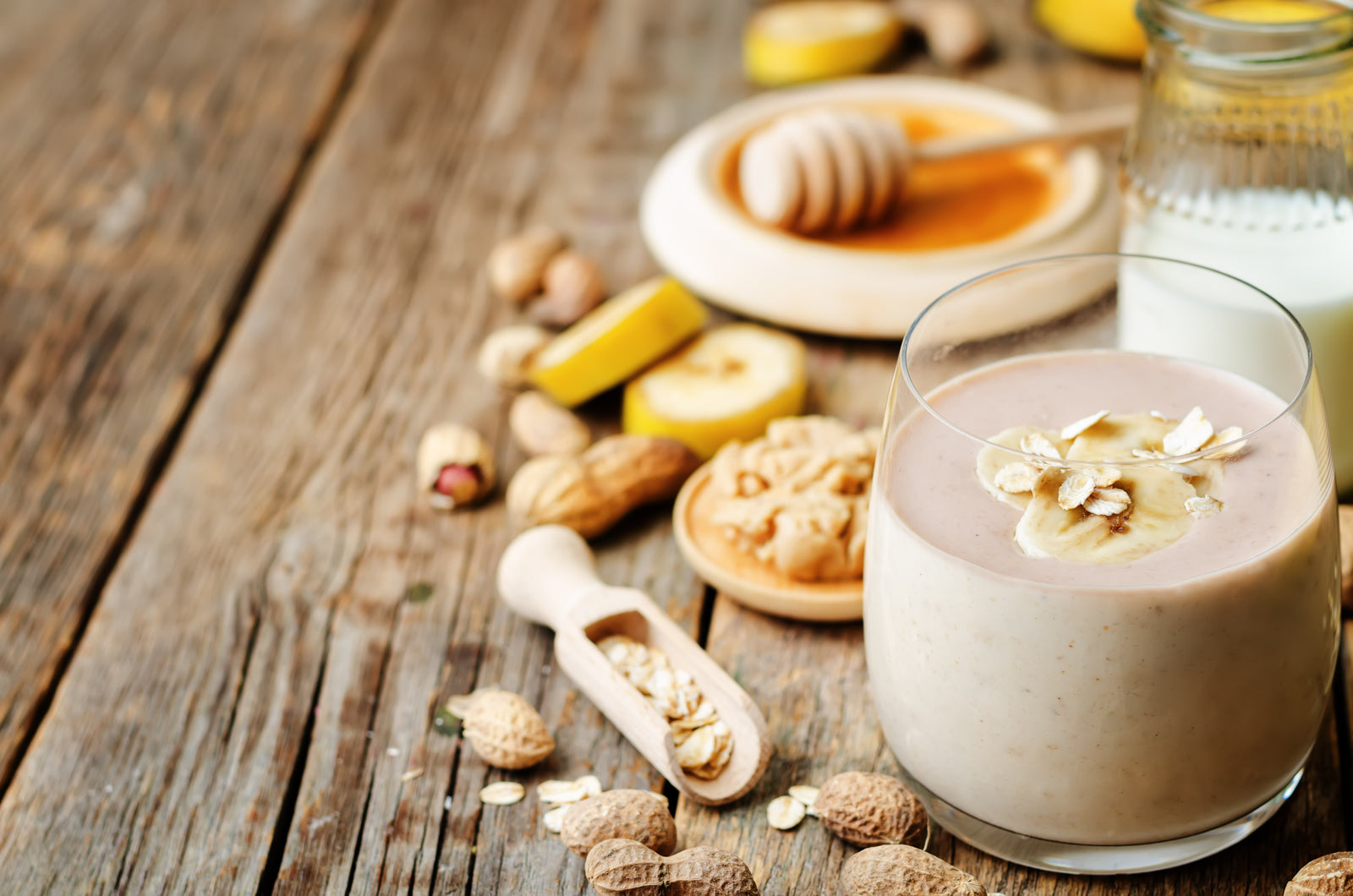 a glass of peanut butter oat proteini smoothie topped with sliced banana and oat surrounded by peanuts, sliced banana, honey on the wooden plate, and a half-full jar of milk