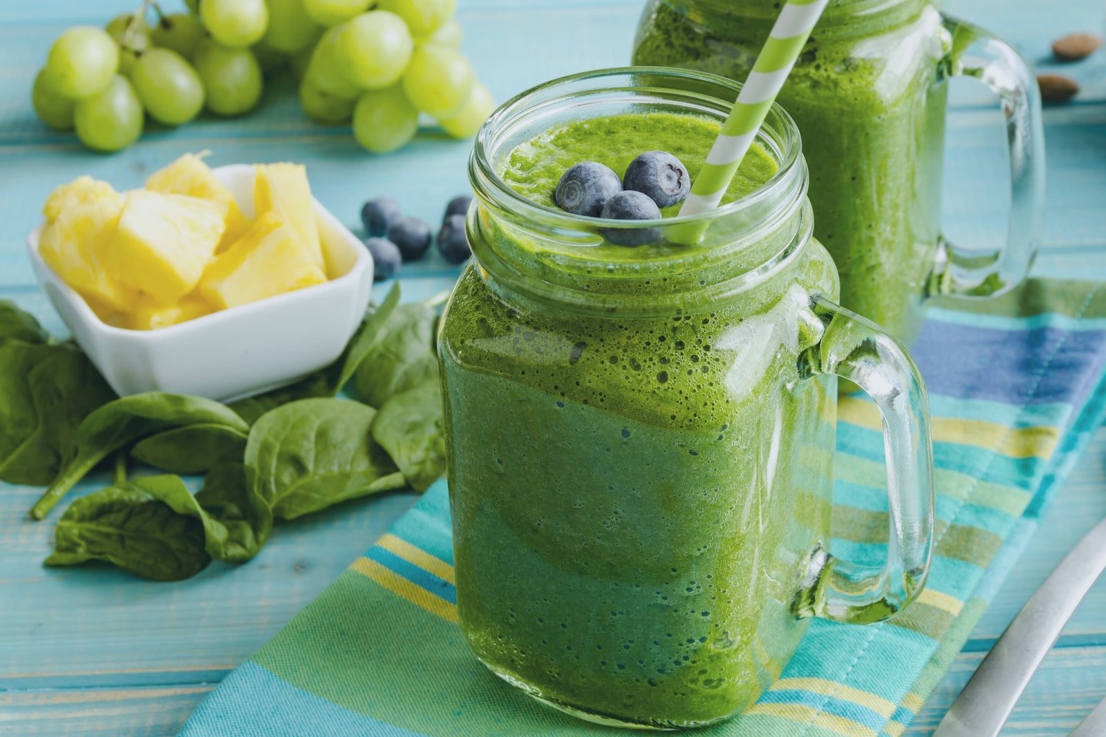 side view of two mason jars filled with pressed greens blueberry lemon smoothie topped with berries on top of a stripe folded table cloth surrounded by a bunch of spinach, a small bowl of slices of pineapple, and grapes on top of a table