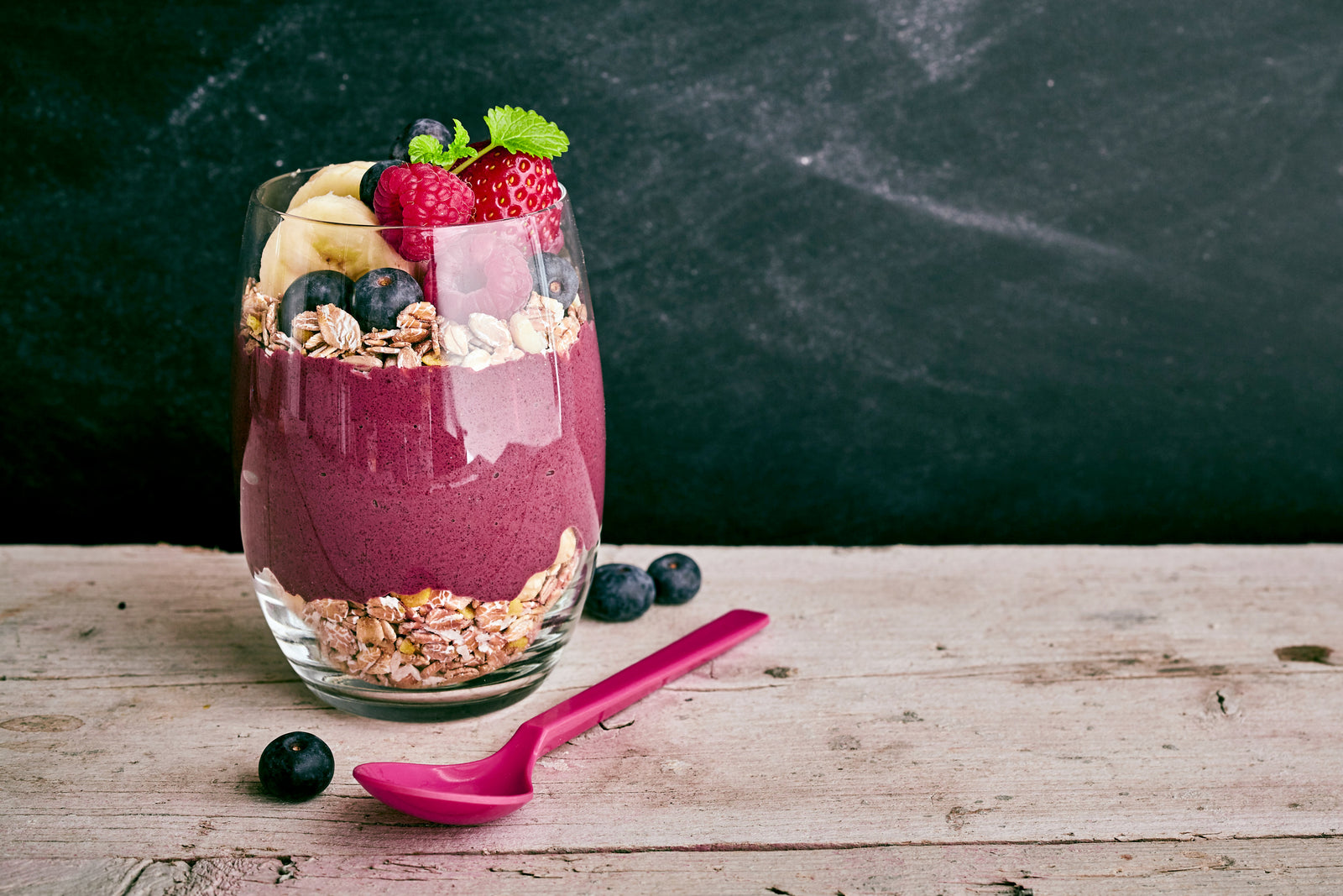1 glass of berry smoothie- on the bottom is oats, in the middle is the smoothie and then topped with more oats, 2 slices of banana, blueberries, and strawberries. On the surface is a pink plastic spoon and 3 blueberries.