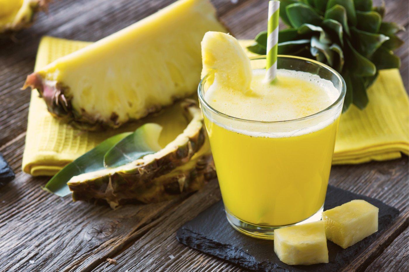 a glass of pineapple chia immunity wellness smoothie garnished with a slice of pineapple with a yellow-green patterned paper straw with two slices of pineapple on top of a black-painted square wood surrounded by pineapple peel and a wedge of pineapple