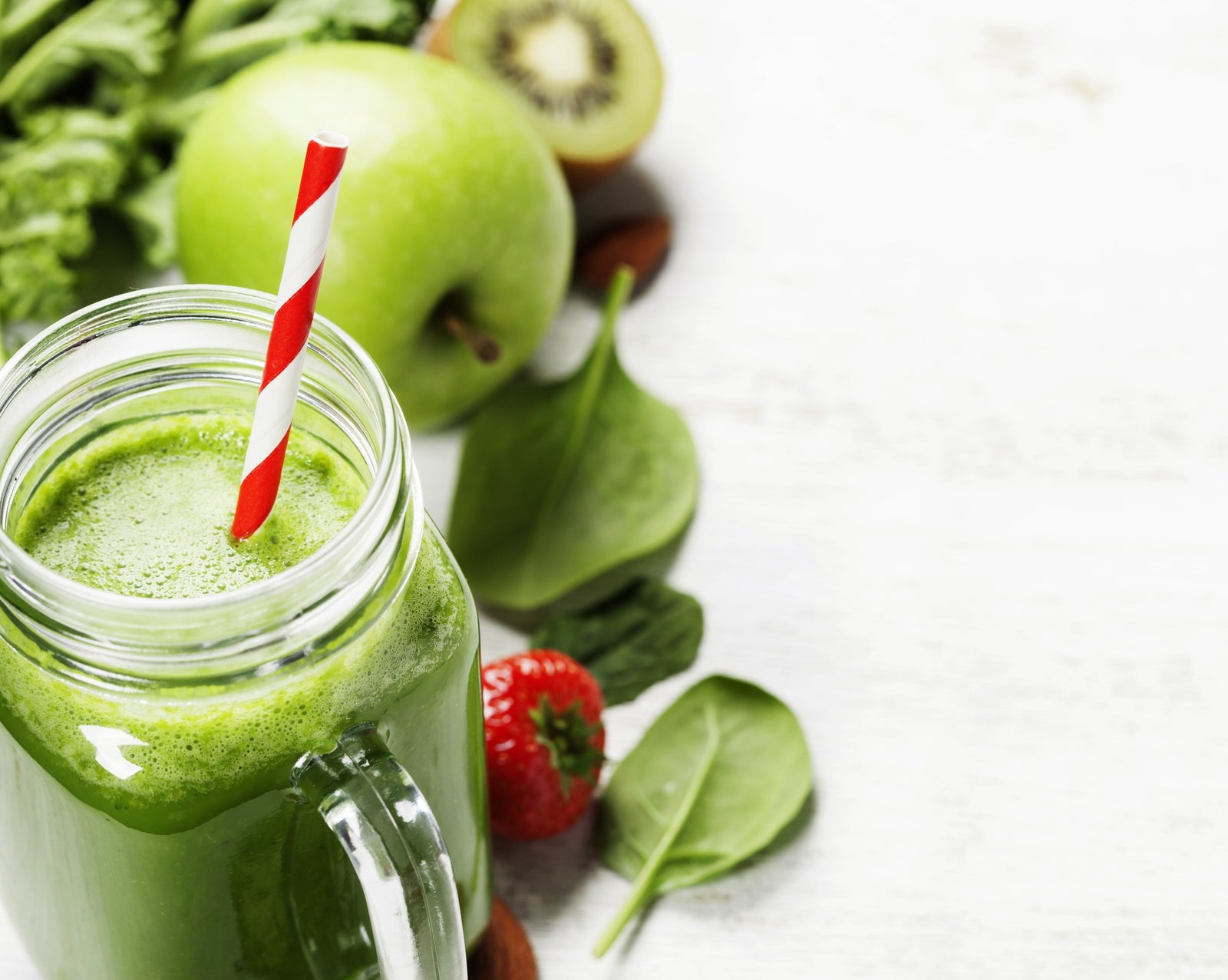 a jar of kale pineapple chia mojito with red patterned paper straw surrounded by spinach, strawberries, an apple, a half-slice of kiwi on top of a white-painted table