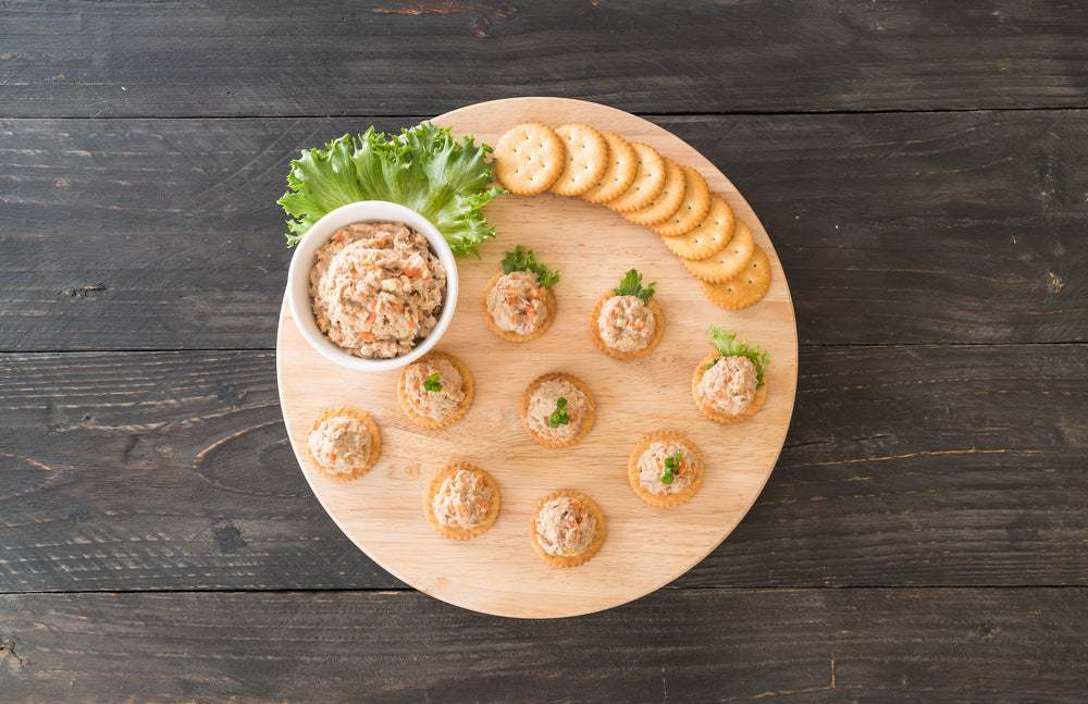 top view of a not tuna-tuna salad with a small bowl of tuna salad with a leaf of kale and biscuits on the side on top of a small circle-shaped wooden table on top of a wooden black table