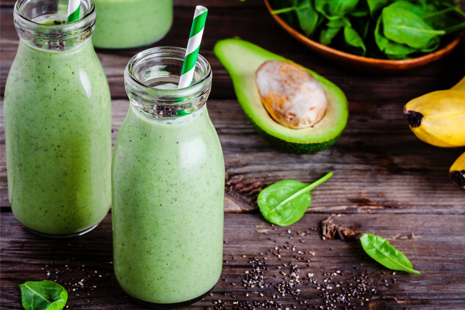 two jars of spa greens topped with chia seeds with green patterned paper straw surrounded by a bowl of arugula, a half-slice avocado, a banana on top of a wooden table