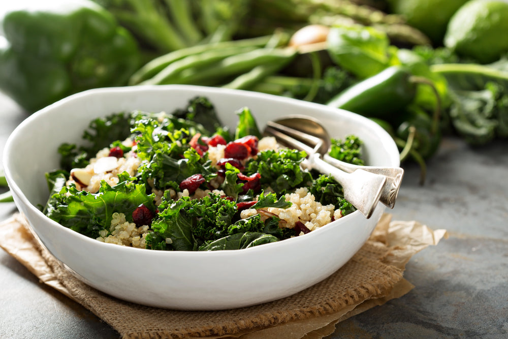 side view of a white bowl with kale Waldorf salad with spoon and fork on top of a weaved cloth with paper on the bottom with green fresh vegetables on the side of the bowl