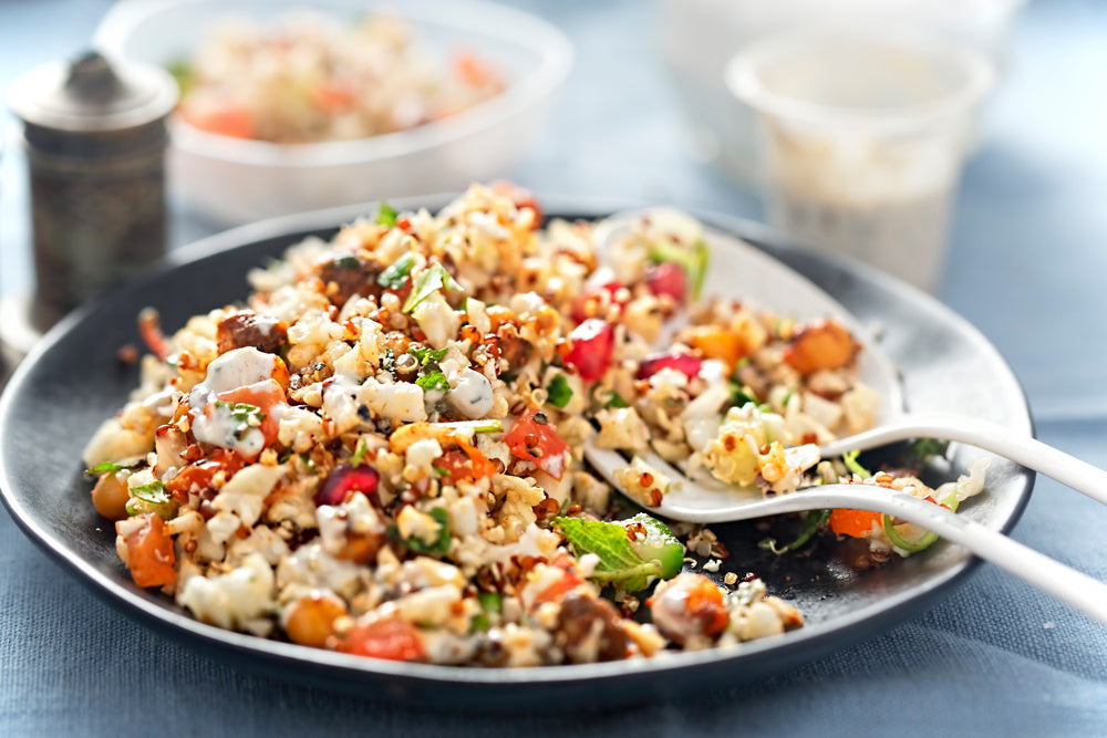 a plate of turmeric cauliflower with quinoa & red beans with stainless spoon and fork at the top with a white plastic cup and a plastic bowl of cauliflower with quinoa & red beans at the back