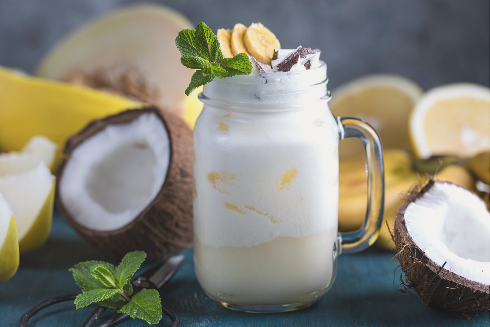 a jar of pina colada smoothie topped with slices of ripe banana with mint and coconut surrounded by coconut, slices of pears, wedges of lemon, a slice of melon, and a banana with the scissor on the side with mint leaves on top on the top table