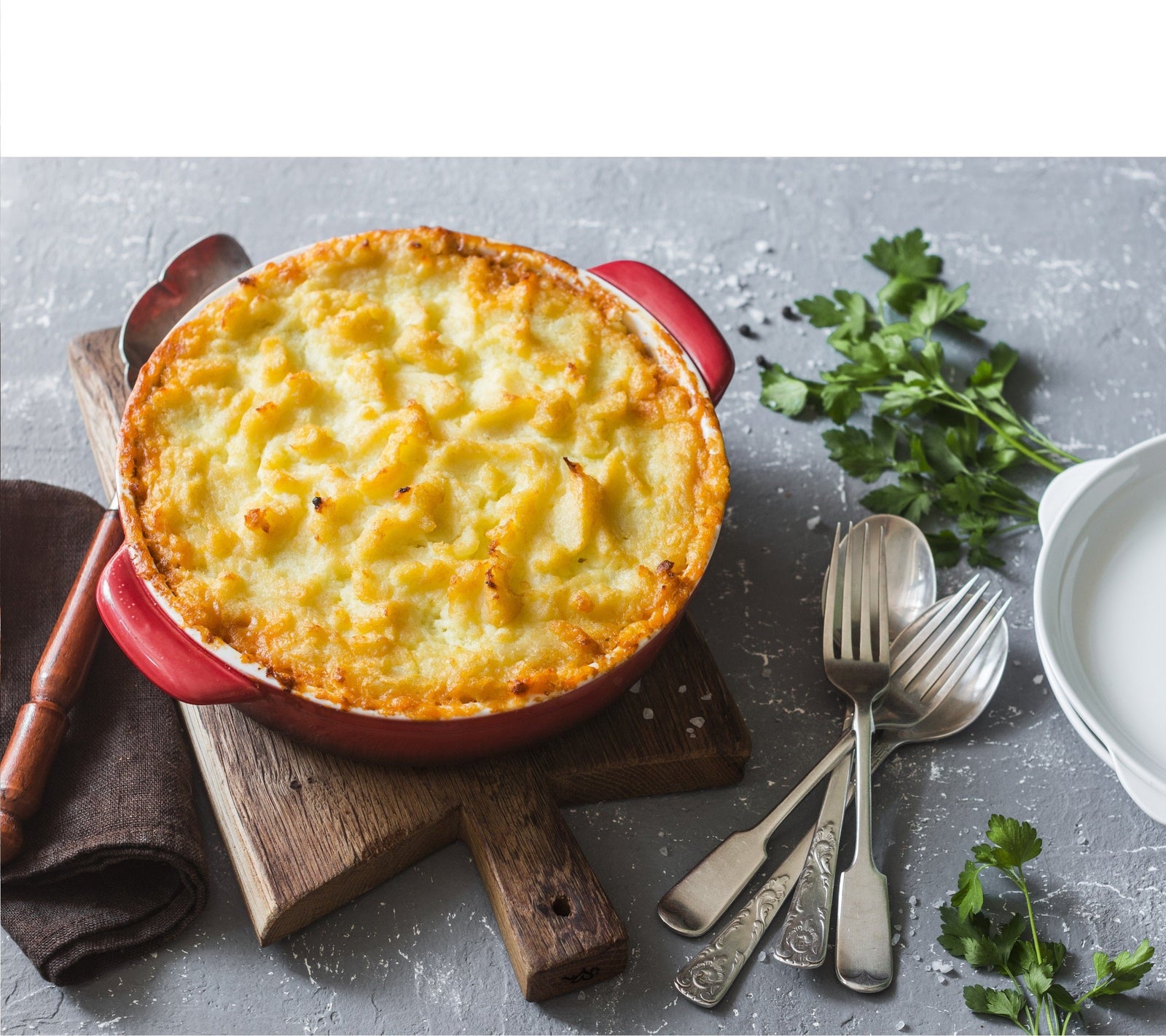 1 pot of Jamaican Yuca Shepherd's Vegan Pie on a wooden chopping board with a spatula and a brown napkin on the left side and a spoon and fork, cilantro, and white plates on the right side.