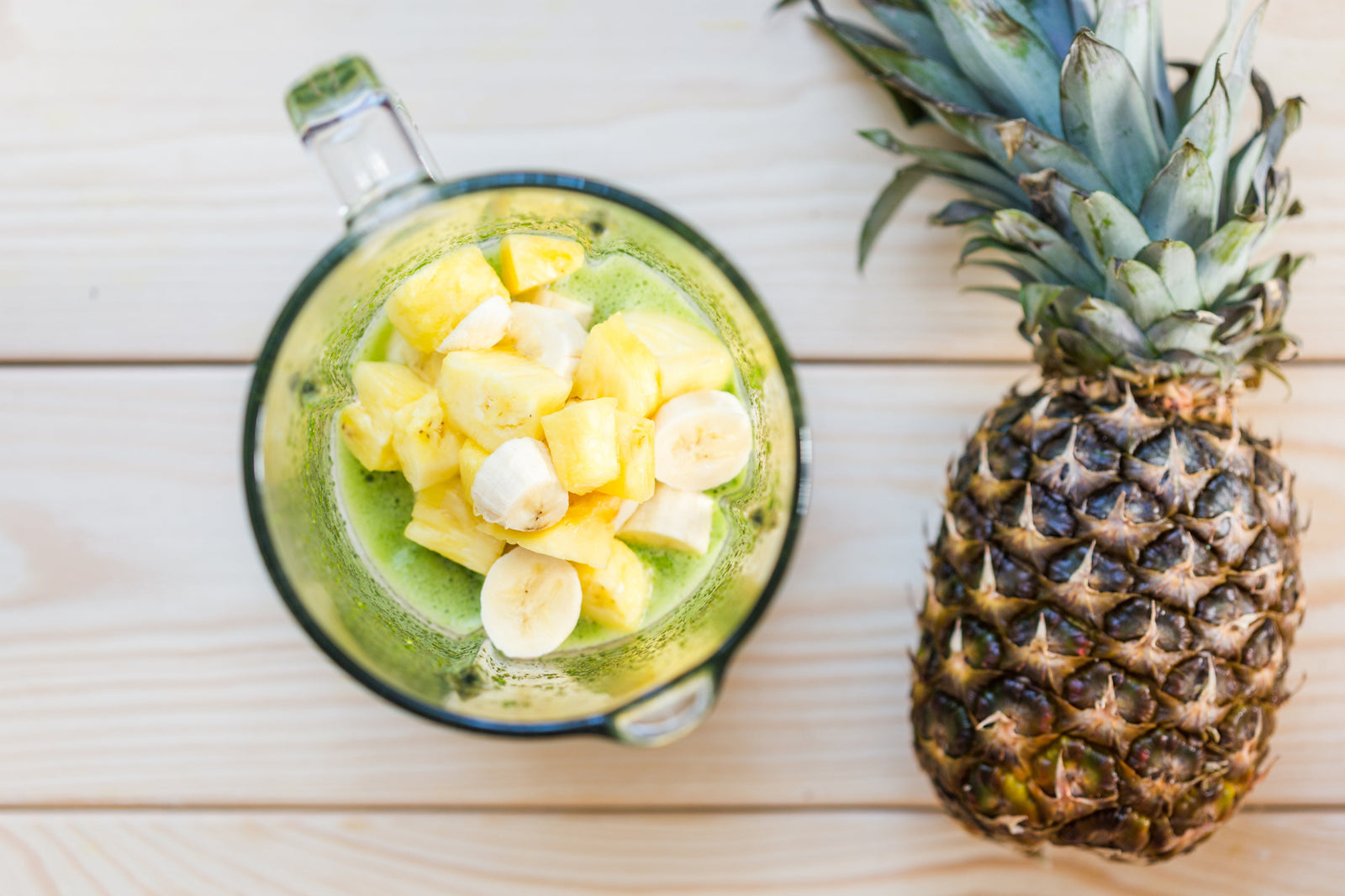 top view of a pitcher with slices of banana, mango, and pineapple with the whole pineapple on the side on top of a wooden table