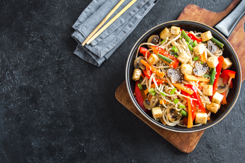 top view of a skillet on a wooden chopping board with a folded blue table clothe on the side with chopsticks on the top filled with szechuan tempeh and broccoli