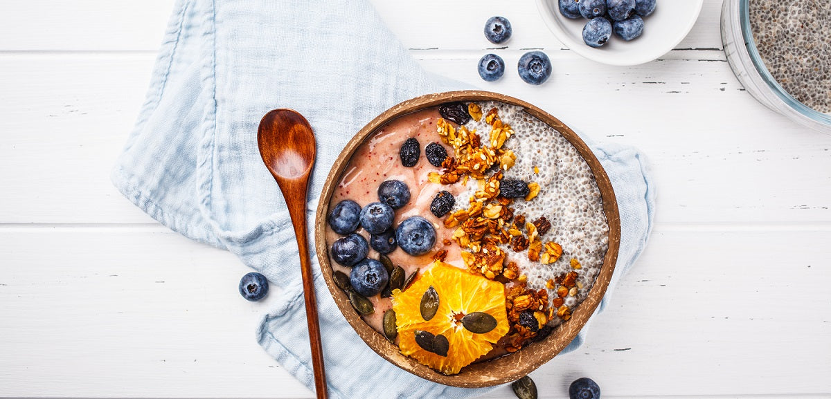 top view of a wooden bowl filled with signature revive beets + roots smoothie topped with chia seeds, berries, sliced pineapple, and nuts on top of a white table cloth with a wooden spoon on the side surrounded by berries and chia seeds