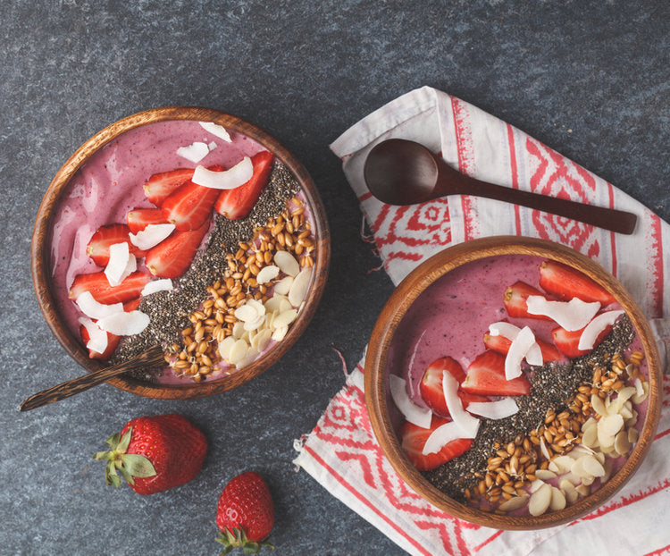 top view of two bowls of berry bliss bowl topped with slices of strawberries, coconut chips, chia seeds and nuts on top with folded tribal red patterned table clothe with a wooden spoon and two strawberries on the side.
