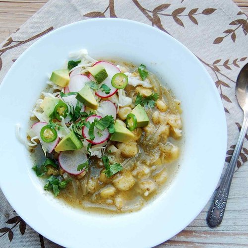 top view shot of a bowl Vegan Pozole Verde- a soup with chickpeas, garnished with chopped avocado, slices of radish, chopped parsley, and chopped green bird chili.