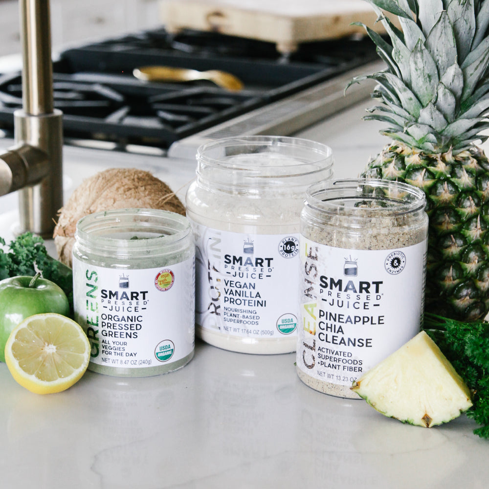 One 240 grams open jar of Organic Pressed Greens, one 500 grams open jar of Vegan Vanilla Proteini, and one 375 grams open jar of Pineapple Cleanse side by side surrounded by a slice of pineapple, parsley, whole pineapple, 1 coconut, 1 apple, and half an orange in a kitchen setting.