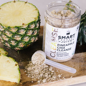 Half pineapple beside an open jar of 375 grams Pineapple Chia Cleanse and in front is a scoop full of light brown powder on a wooden surface.