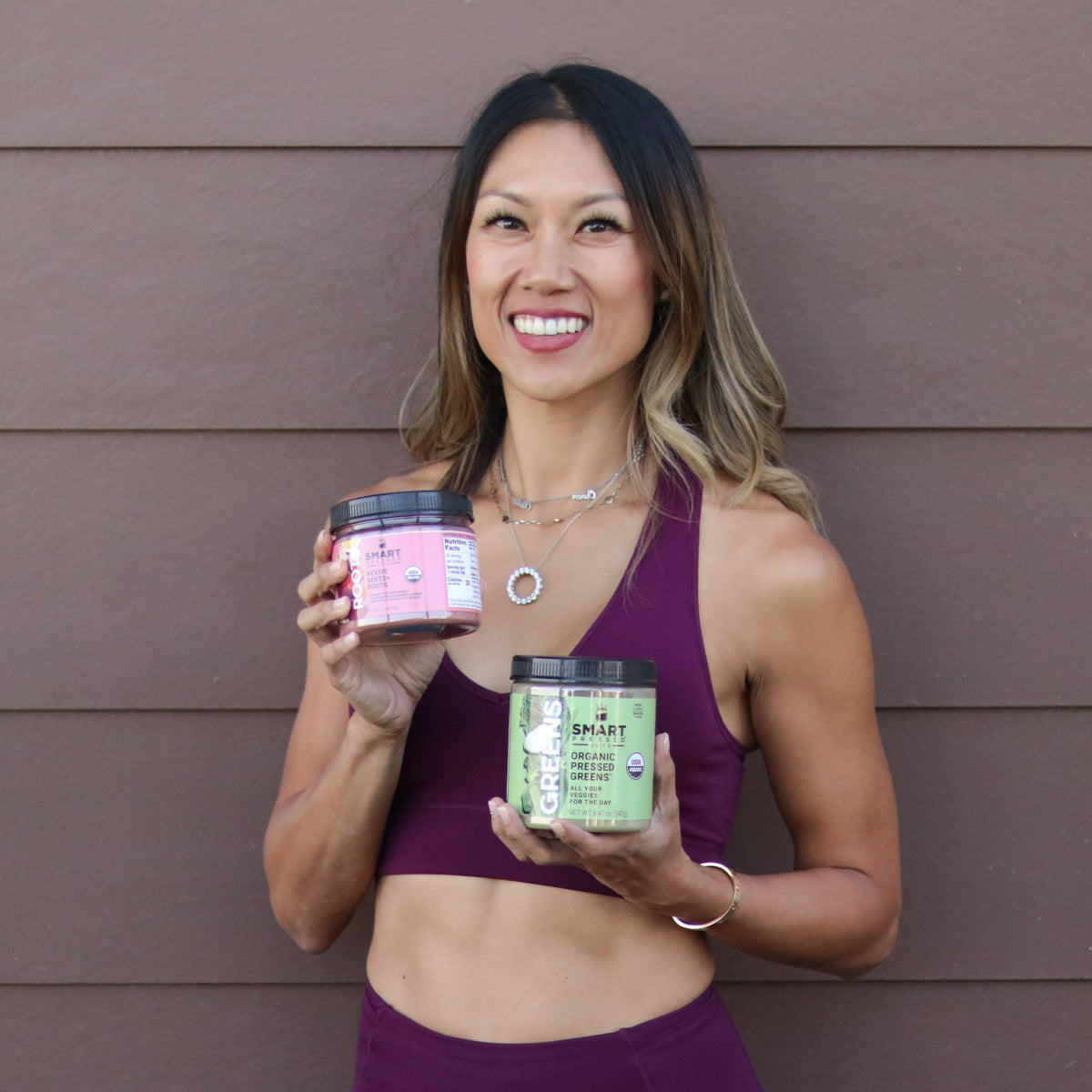 A beautiful and healthy-looking woman holding 1 jar of Revive Beet+Roots on her right hand and 1 jar of Organic Pressed Greens on her left hand. She is standing against a brown wall.