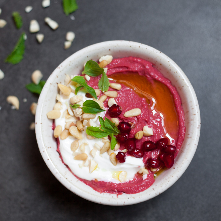 a bowl of Revive Beet+Roots hummus topped with olive oil, coconut whipping cream, pomegranate seeds and garnished with green herbs and nuts. 