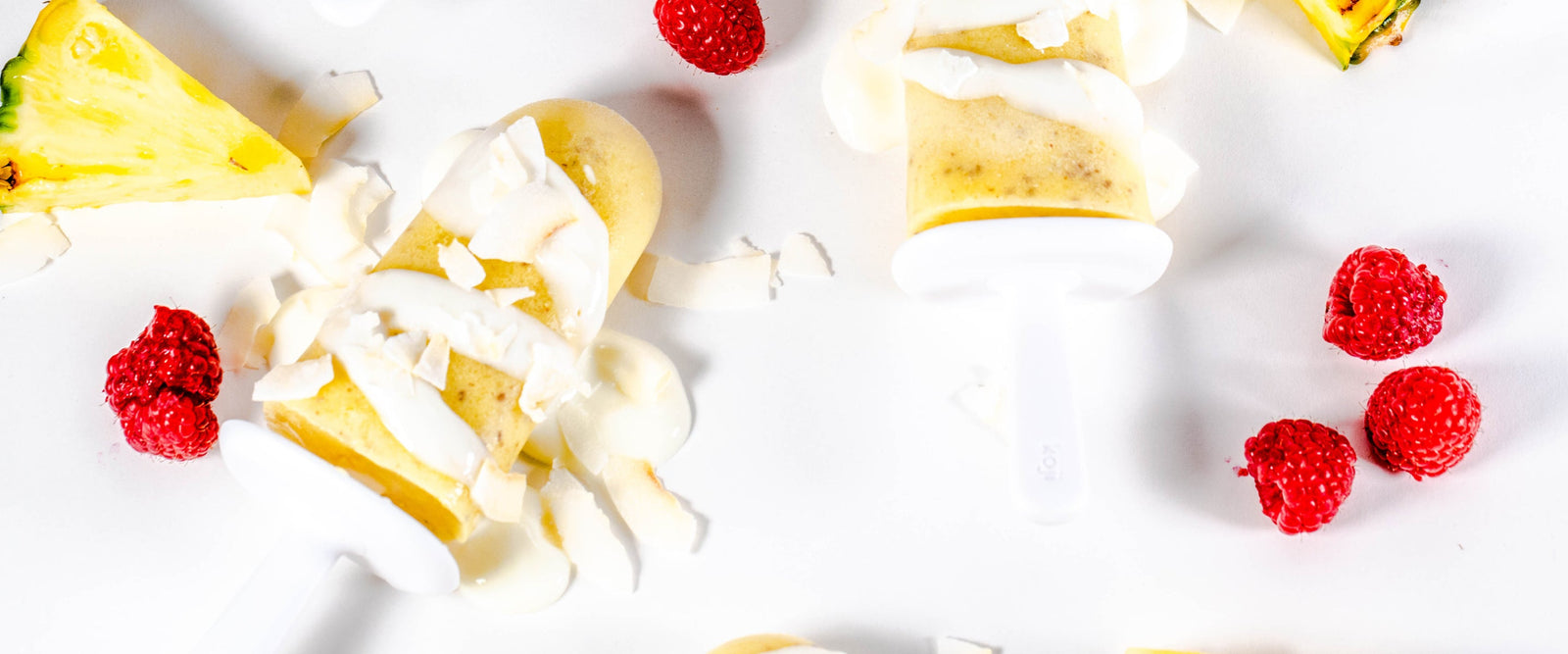 top view of two pineapple popsicles with chia seeds top with coconut bits surrounded by berries and a slice of pineapple on top of a white table