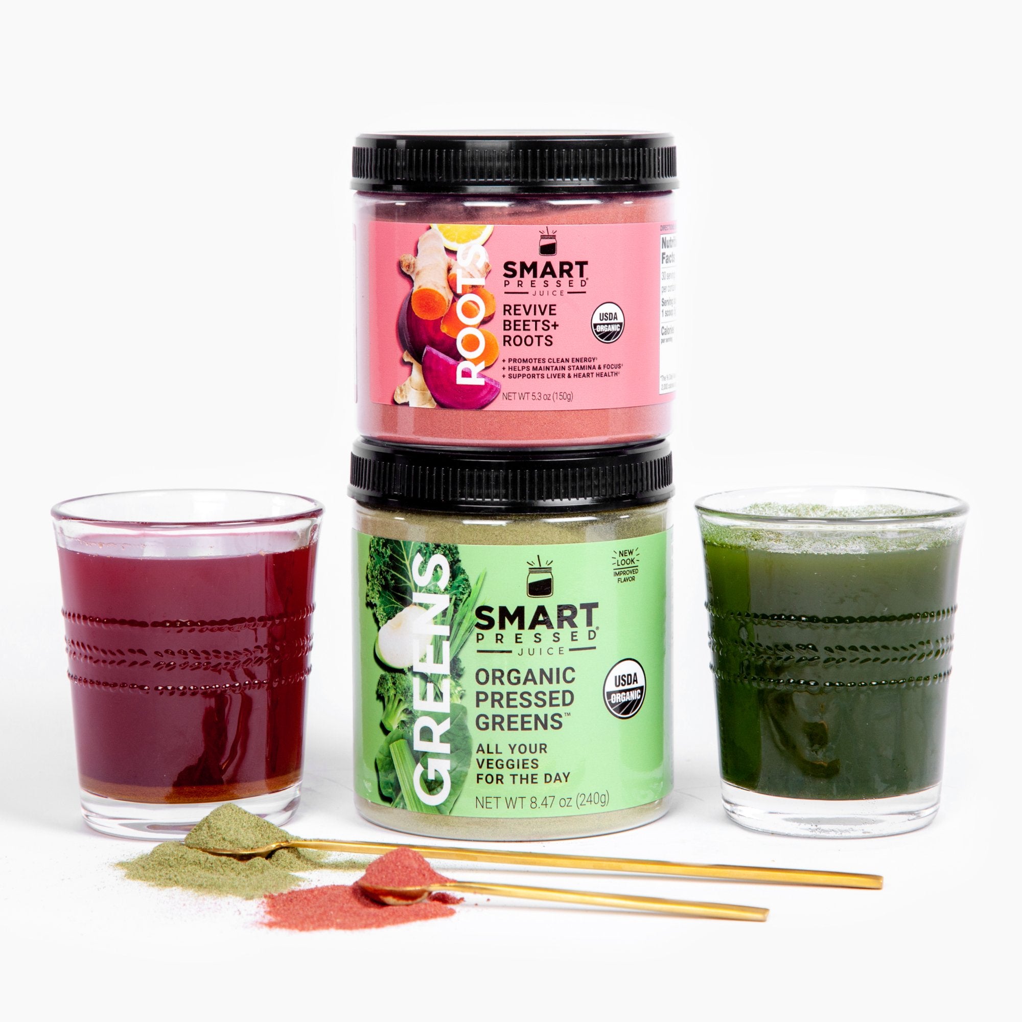 Image of a glass with a purple juice beside a jar of Revive Beet+Roots stacked in a jar of Organic Pressed Greens, and beside is a glass with a green juice. In front is 2 stirring spoons full of green and dark pink powder. Set against a white background.